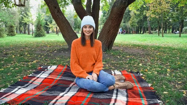 happy stylish woman smiling at camera while sitting on plaid blanket in park - Photo, Image