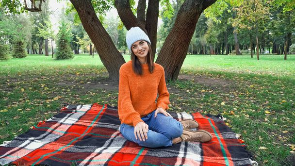 joyful woman smiling at camera while resting on checkered blanket in autumn park - Photo, image