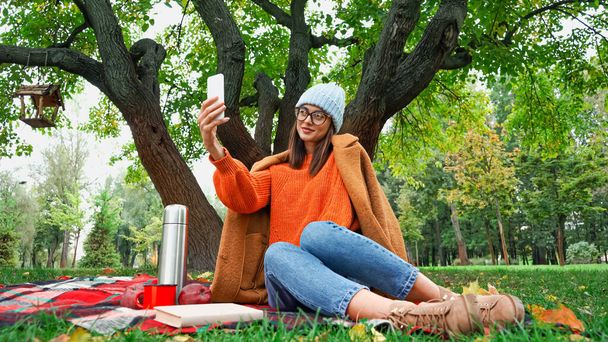 χαμογελαστή γυναίκα σε κομψό φθινοπωρινό ντύσιμο λήψη selfie στο κινητό τηλέφωνο κατά τη διάρκεια πικνίκ στο πάρκο - Φωτογραφία, εικόνα