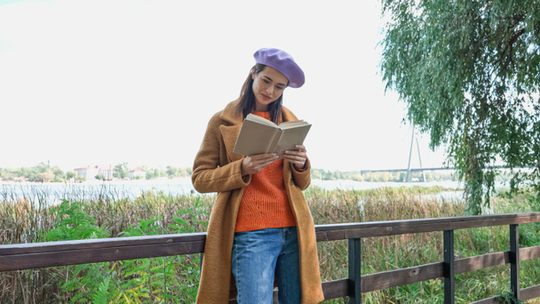 young woman in trendy autumn clothes reading novel on bridge in park - Photo, Image