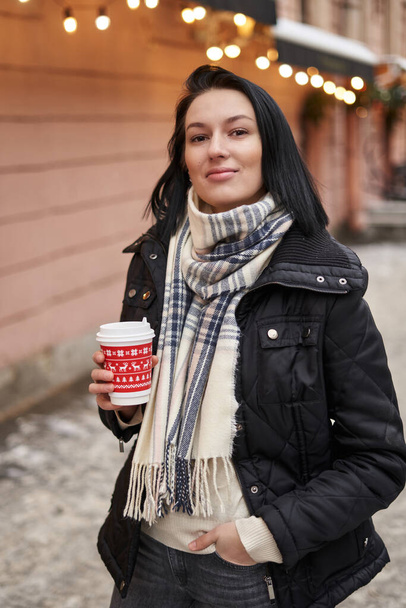 a beautiful girl with shoulder-length dark hair stands on the street and holds a red glass with a drink in her hands. bokeh blurred lights in the background                                - Photo, Image