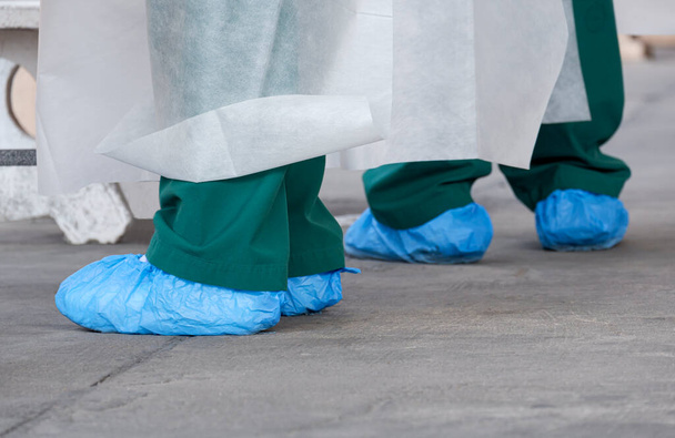 Nahaufnahme der Füße des Gesundheitspersonals in PSA-Kleidung und blauen Plastiktüten auf dem Boden während der Arbeit im öffentlichen Außenbereich - Foto, Bild