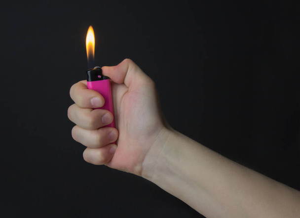 A woman's hand holds a pink lighter. A burning flame from a lighter. Right hand. Close-up, black background - Photo, image