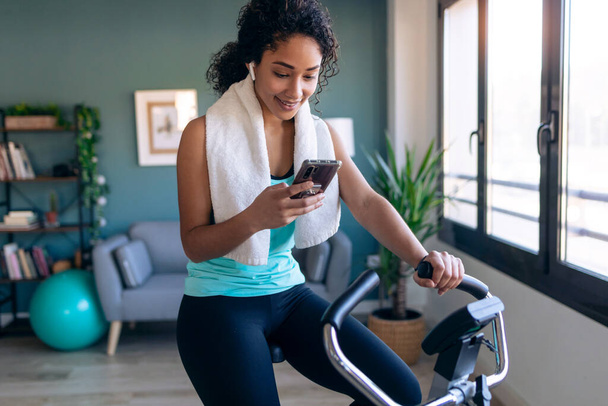 Shot of afro young fitness girl using mobile phone while training on exercise bike at home. - Foto, Imagen