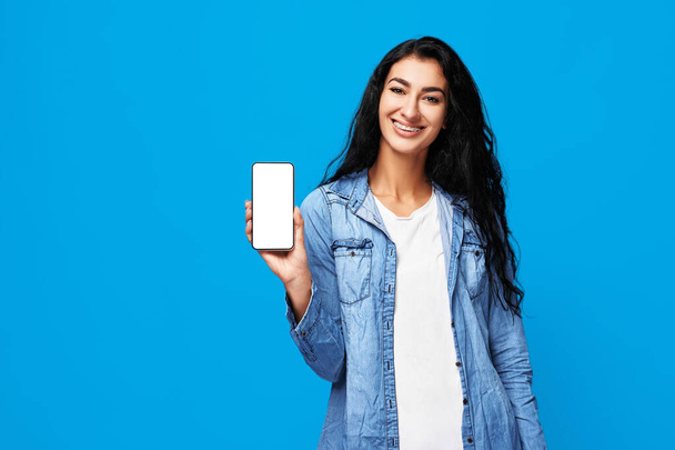 Jeune femme arabe attrayante debout avec un téléphone intelligent dans les mains. Isolé en studio sur fond bleu. - Photo, image