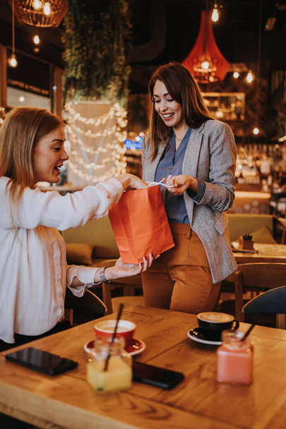 Two best friends meet in a nice restaurant or coffee bar. One prepares a surprise for the other and gives her a birthday present. Happy young people.  - Foto, imagen