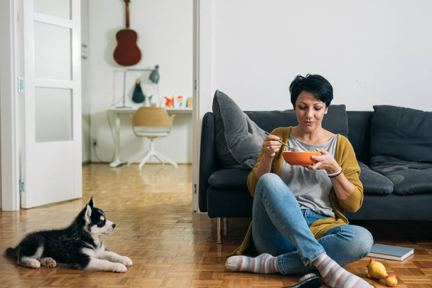 woman eating snack, sitting on the floor in her living room, her dog is beside her - Foto, afbeelding