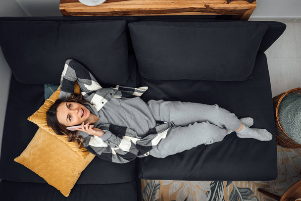 Top view of an attractive girl lying on a sofa, wearing home cloths with no shoes and a cell phone on her ear. Relax talking on the couch in the living room. Chatting with friends, dating on phone. - Photo, Image