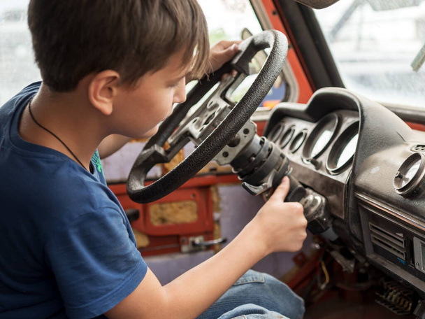 An 11-year-old boy enthusiastically plays the driver behind the wheel of an old abandoned car. - Zdjęcie, obraz