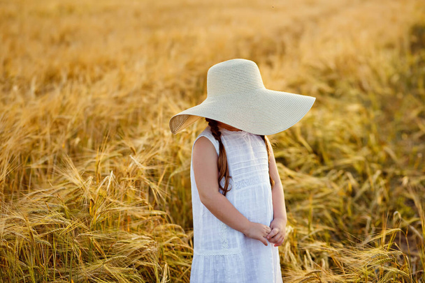 Portrait of a beautiful little girl in a white sundress and a large straw hat covering her face on a golden wheat field at sunset. - Fotó, kép