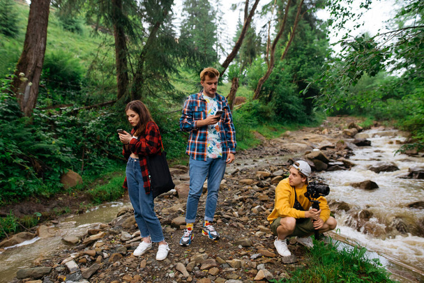 Les jeunes dans les montagnes près du ruisseau se reposent sur une randonnée, un gars avec un stabilisateur est assis sur le sol et des amis utilisent un smartphone. Hipsters lors d'une randonnée en montagne. - Photo, image