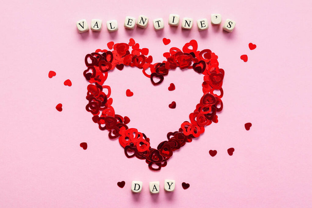 Wooden cubes with the words Valentine's Day and heart made by heart shaped confetti on pink background, top view. Valentine's Day card with wooden letter blocks. - Zdjęcie, obraz