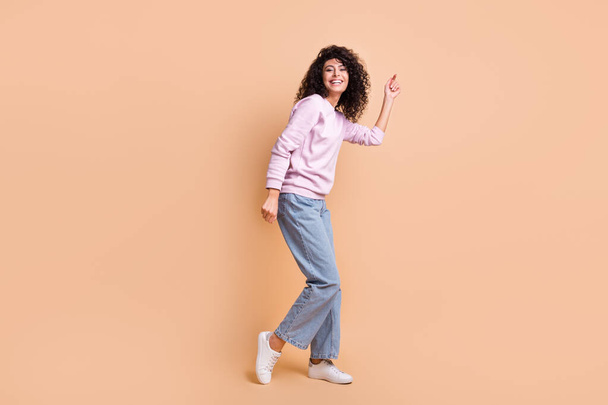 Full size profiel foto van optimistisch krullend meisje dansen slijtage trui jeans sneakers geïsoleerd op perzik kleur achtergrond - Foto, afbeelding
