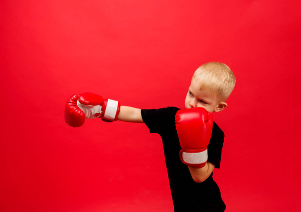 ένα μικρό αγόρι πυγμάχος με κόκκινα γάντια πυγμαχίας στέκεται κάνοντας μια γροθιά σε ένα κόκκινο φόντο με χώρο για κείμενο - Φωτογραφία, εικόνα