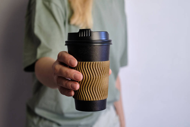 Woman holding in hand black paper cup with coffee or tea. Drink coffee time. Disposable paper cup. Beautiful blond hair young girl. Casual style, shirt and jeans. Take away. Hot beverage. Blank space - Фото, изображение