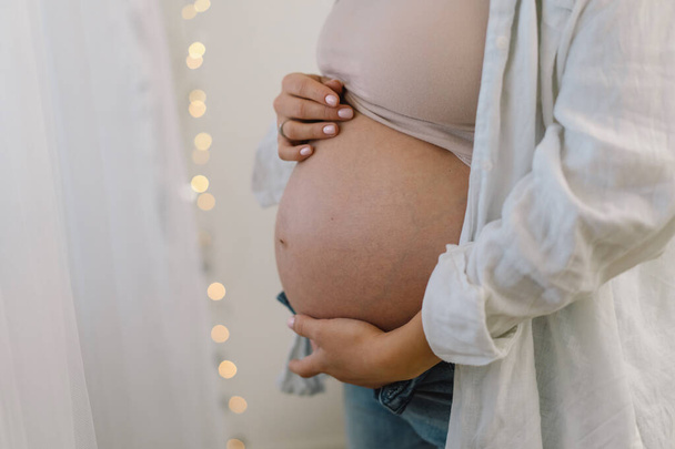 魅力的な妊婦は窓のそばに立って腹を抱えている。妊娠の最後の数ヶ月 - 写真・画像