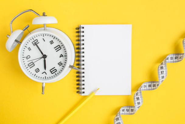 Top view of open notepad, white alarm clock and white centimeter on the yellow  background - Fotoğraf, Görsel