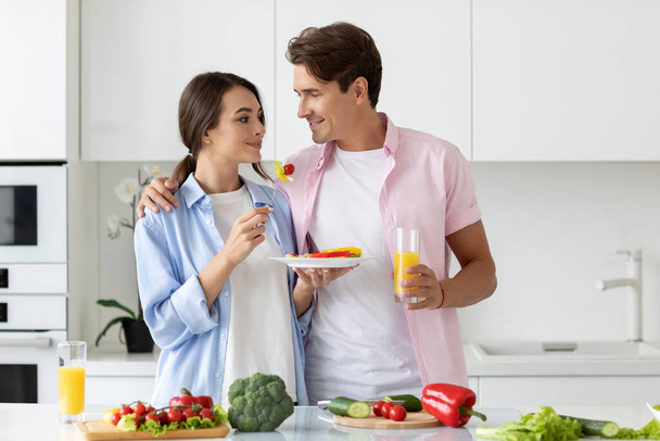 Smiling woman feeding her man tasting healthy vegetable salad in the kitchen. Happy family eat at home. - Photo, Image