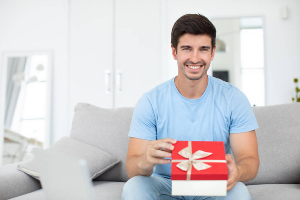 Young handsome smiling man with a gift in his hands sits on the sofa at home. - Foto, Imagem