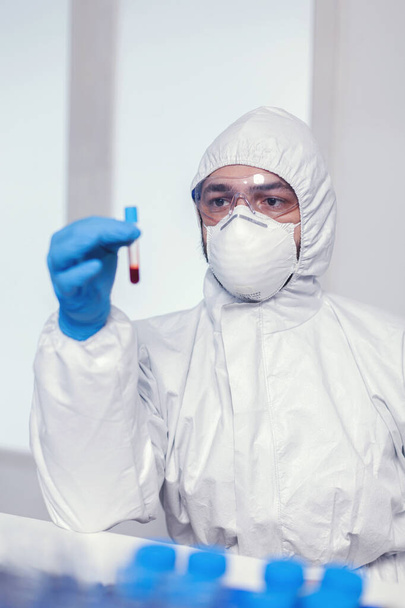 Bioquímico en traje de ppe buscando y trabajando con sangre en tubo de ensayo - Foto, imagen