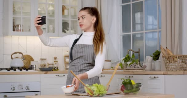 Jonge vrouwelijke chef-kok opnemen video recept op smartphone, met verse salade ingrediënten om de camera in de keuken - Video