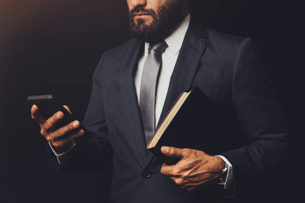 hombre de traje sosteniendo una Biblia en su brazo y con el teléfono celular en la mano - Foto, Imagen