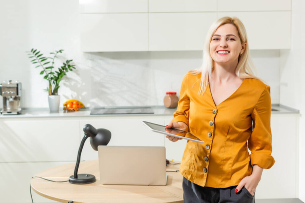 Portret van een vrouw die met een laptop werkt in een lichte keuken. Covid-19 coronavirus. Sociale afstand - Foto, afbeelding