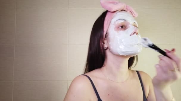 Morena jovem com uma faixa de cabeça rosa aplicando máscara de alginato no rosto usando escova cosmética. Procedimentos de cuidados com a pele em casa. - Filmagem, Vídeo