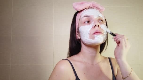 Молода брюнетка з рожевою пов'язкою наносить маску алкінату на обличчя за допомогою косметичної щітки. Процедури догляду за шкірою вдома
. - Кадри, відео