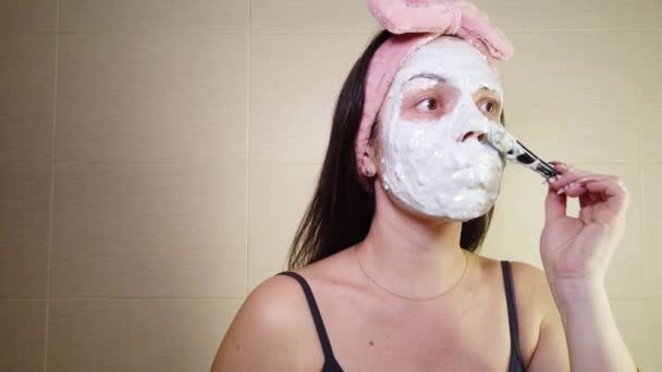 Giovane bruna con un cerchietto rosa che applica una maschera di alginato sul viso usando un pennello cosmetico. Procedure di cura della pelle a casa. - Filmati, video