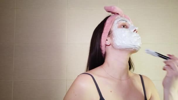 Mladá brunetka s růžovou čelenkou nanáší alginátovou masku na obličej pomocí kosmetického kartáče. Procedury péče o pleť doma. - Záběry, video