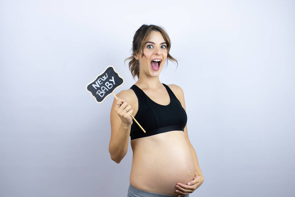 Beyaz arka plan üzerine spor giysili hamile genç bir bayan hayrete düştü ve elinde yeni bebek mesajıyla kara tahtaya sarıldı. - Fotoğraf, Görsel