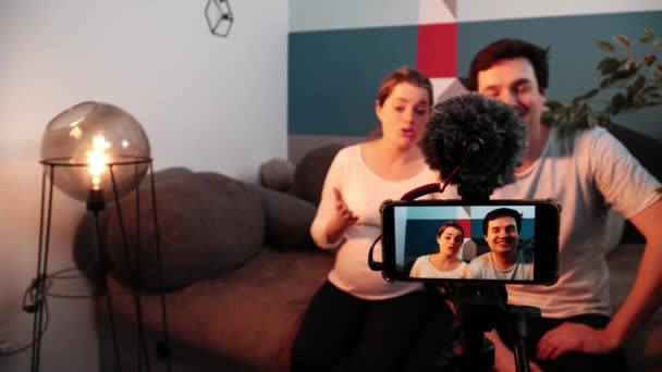 Mężczyzna i kobieta w ciąży udzielają wywiadu siedząc na kanapie w salonie przed kamerą mobilną - Materiał filmowy, wideo