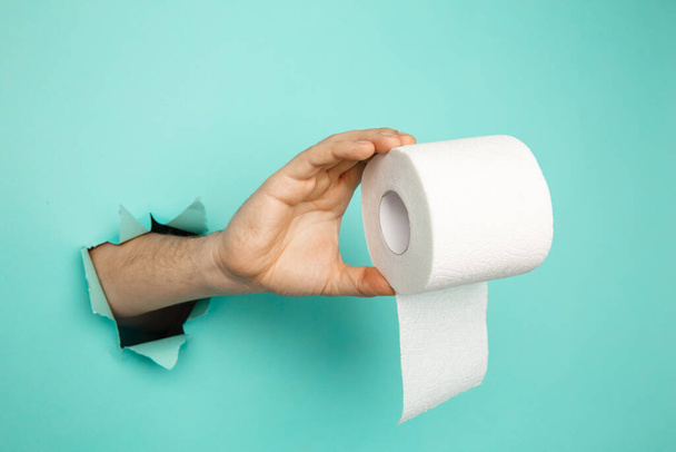 Mans hand holding a roll of toilet paper from blue torn background - Photo, Image