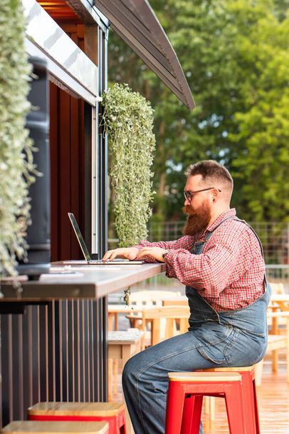 Portrait of brutal bearded hipster man freelancer wearing blue overalls and checked shirt working on laptop sitting in cafe/restaurant outdoors. Side view. Distance job, remote work. - Photo, Image