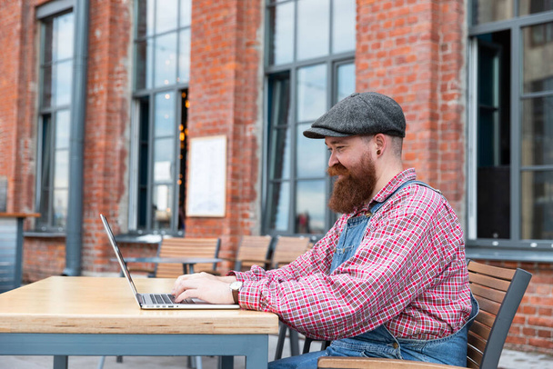 Ritratto di brutale hipster barbuto freelance che indossa tuta blu e camicia a quadri che lavora su laptop seduto in un bar / ristorante all'aperto. Vista laterale. Lavoro a distanza, lavoro a distanza. - Foto, immagini