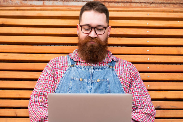 Close up portrait of serious brutal bearded hipster man in blue overalls, checked shirt working on laptop sitting on bench outdoors. Distance job, remote work. - Fotografie, Obrázek