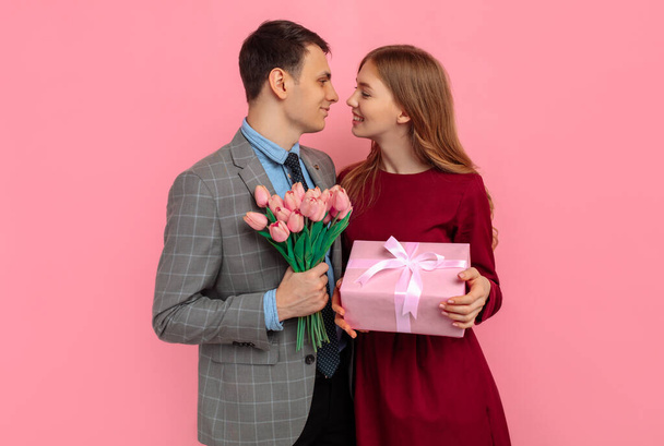 Handsome elegant man in classic suit giving flowers and gift box to his beautiful girlfriend on isolated pink background, Valentine's Day concept - Photo, image