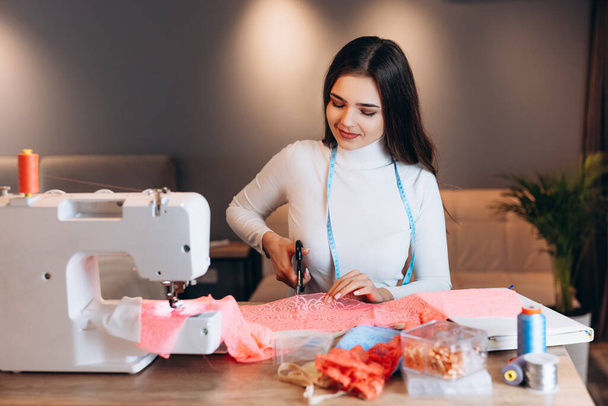 Mujer modista joven cose ropa en la máquina de coser. Usando tijeras para cortar textiles en taller. Creación de cursos de diseño de ropa en línea. - Foto, Imagen