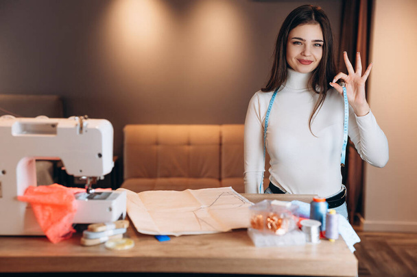 Mujer bonita modista en gafas cose ropa en la máquina de coser. Sonriendo mostrando OK costurera en el taller. Creación de cursos de diseño de ropa en línea. - Foto, imagen