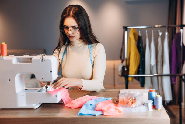 Mooie naaister vrouw naait kleren op naaimachine. In proces naaister in de werkplaats. Het creëren van online kleding ontwerp cursussen. - Foto, afbeelding