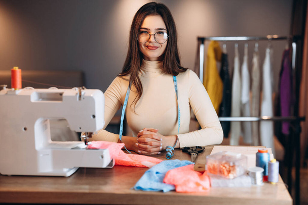 Mujer bonita modista en gafas cose ropa en la máquina de coser. Costurera sonriente en el taller. Creación de cursos de diseño de ropa en línea. - Foto, Imagen