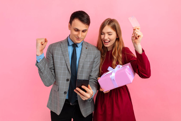 Happy young couple in love, woman and man with gift box, celebrate Valentine's Day, Women's Day, using mobile phone, holding credit card, shopping online, on pink background - Фото, изображение