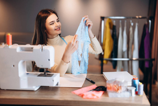 Молодая портниха с швейной машинкой. Швея проверяет текстиль в мастерской. Создание онлайн курсов по дизайну одежды. - Фото, изображение