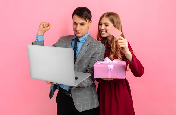 Happy young couple, man and woman with a gift box, celebrating Valentine's Day, Women's Day, shopping online on a laptop, holding a credit card, on a pink background - Foto, Imagem
