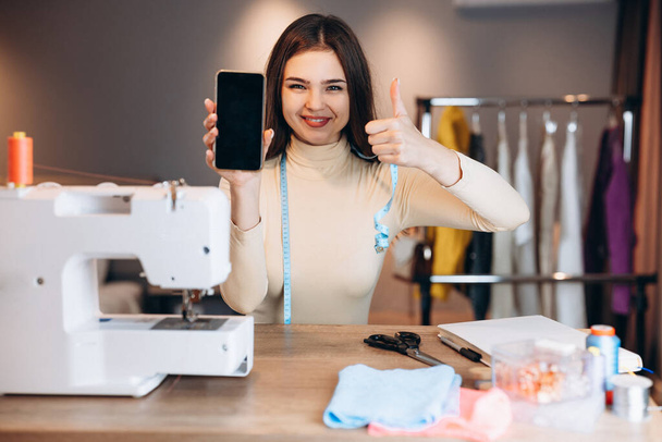 Mujer modista joven cose ropa en la máquina de coser. La costurera muestra felizmente su teléfono en el taller. Creación de cursos de diseño de ropa en línea. - Foto, Imagen