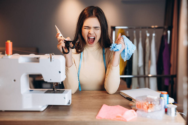 Mujer joven modista con máquina de coser. La costurera está gritando sosteniendo tijeras y textiles en el taller. Creación de cursos de diseño de ropa en línea. - Foto, Imagen