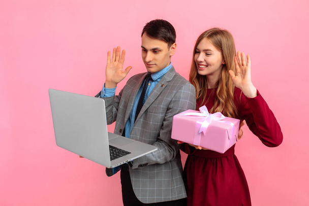 Happy young couple, man and woman with gift box, celebrating Valentine's Day, Women's Day, using laptop computer on pink background - Foto, afbeelding