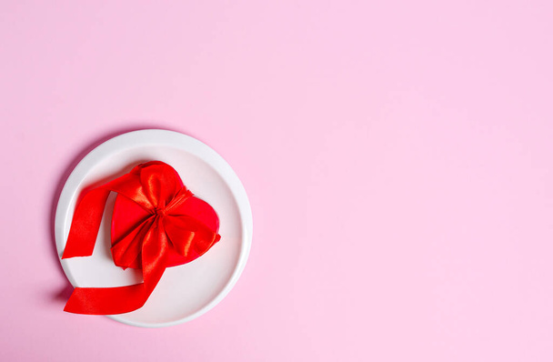 Red gift box in the form of a heart on a white plate on a pink background. Love concept, copy space, flat lay, place for text, banner, top view. - Фото, зображення