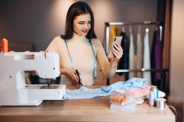 Mujer modista joven cose ropa en la máquina de coser. Mirando costurera teléfono en el taller. Creación de cursos de diseño de ropa en línea. - Foto, Imagen